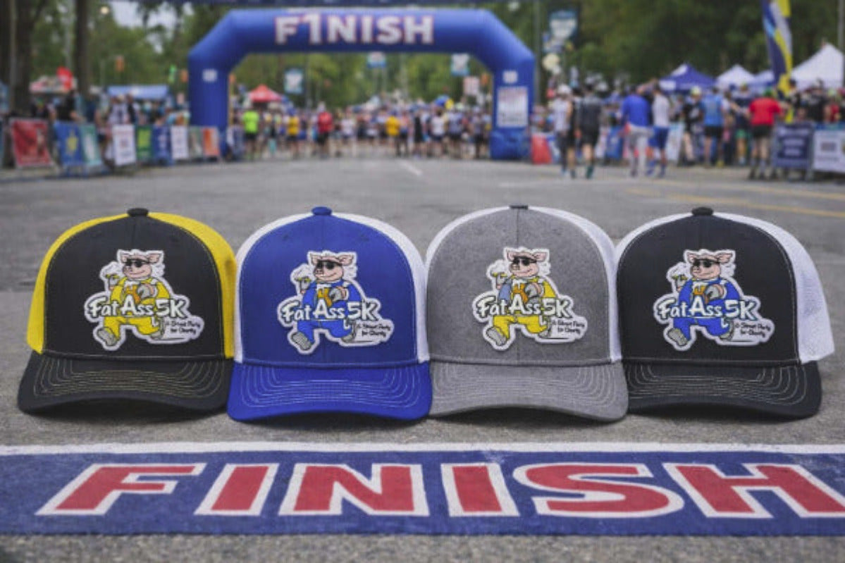 Four hats with a 'Fat Ass 5K' logo on a finish line with race participants in the background.